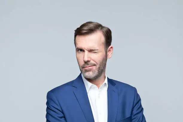 Portrait of mature man wearing navy blue jacket and white shirt, winking at camera. Studio shot of male entrepreneur against grey background.