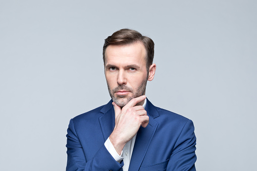 Portrait of mature man wearing navy blue jacket and white shirt, looking at camera with hand on chin. Studio shot of male entrepreneur against grey background.