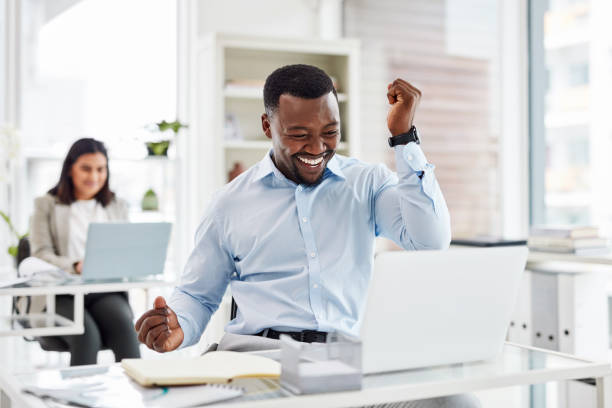 shot of a young businessman cheering while working on a laptop in an office - extatisch stockfoto's en -beelden