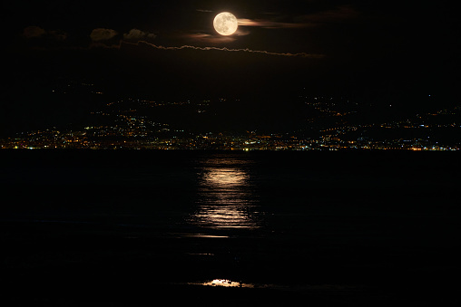 full moon is reflected in the sea and in the background the lights of the  calabrian coast in Italy