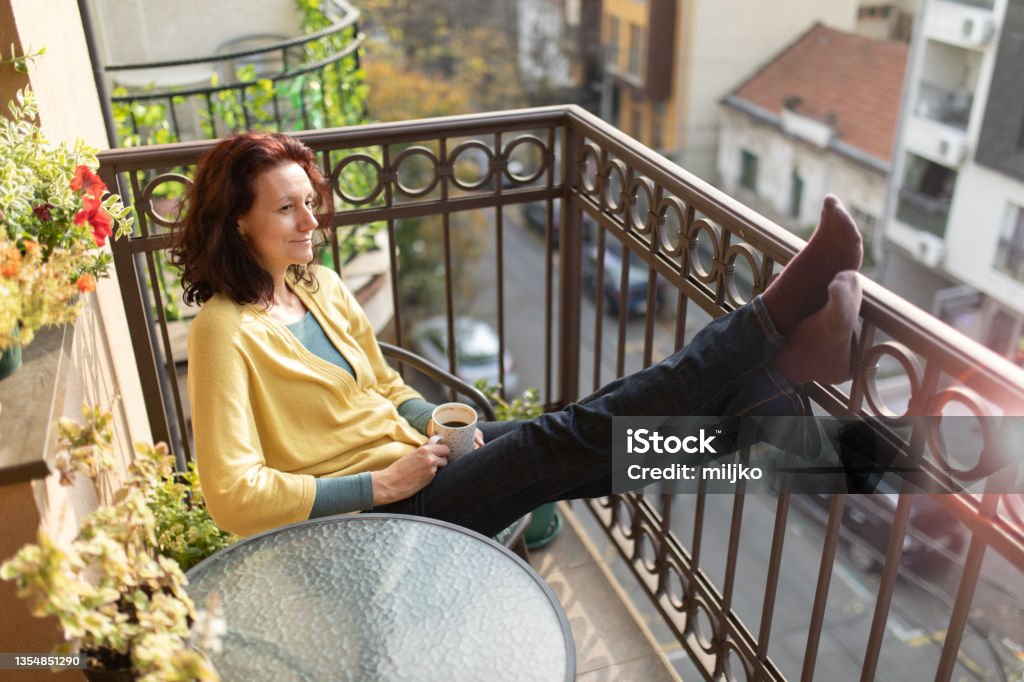 Mature woman relaxing on balcony at morning Beautiful mature woman sitting on terrace, relaxing and enjoying morning sun. She is drinking coffee and enjoying view Balcony Stock Photo