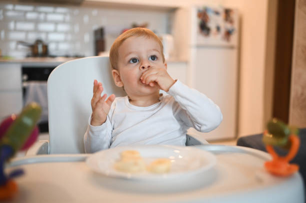 Little happy cute baby toddler boy blonde sitting on baby chair playing with banana. Baby facial expressions indoors at home kitchen interior with food. Healthy eating happy family childhood concept. stock photo