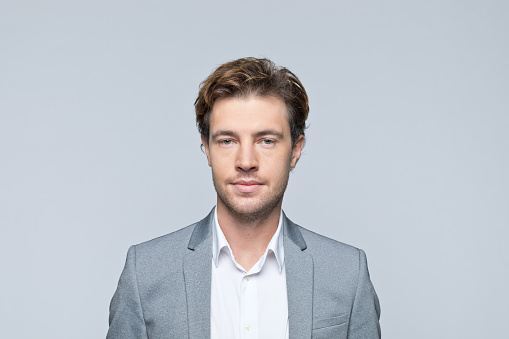 Portrait of handsome young man wearing grey jacket, smiling at camera. Studio shot of male entrepreneur against grey background.