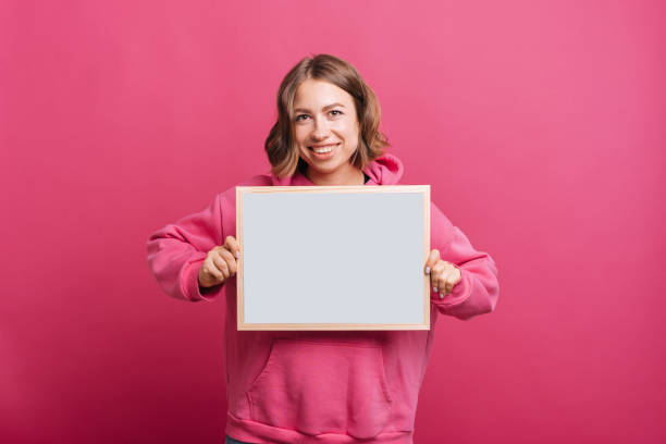 A cheerful young woman is holding a whiteboard while smiling at the camera near a pink wall stock photo