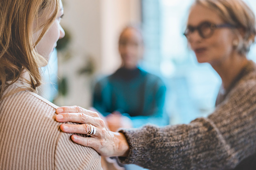 Senior woman comforting a female with hand on her shoulder during group therapy session. Close-up of a woman getting psychological support during therapy session