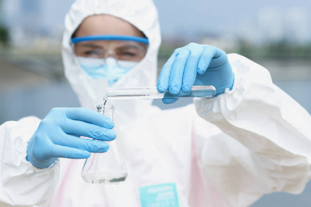 ricercatore in tuta protettiva e occhiali con guanti versa acqua dal becher di vetro della provetta - test tube biologist laboratory beaker foto e immagini stock