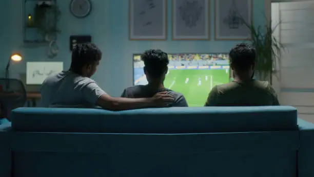 Back view of Indian male friends sitting on couch in living room and talking with each other while watching soccer competition on TV in evening