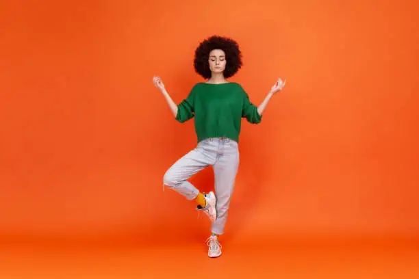 Photo of Full length portrait of woman with Afro hairstyle wearing green sweater with mudra gesture hands up, closed eyes, meditating standing in yoga position.