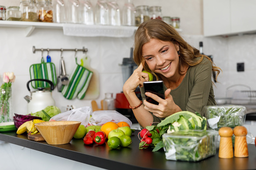 Copy space shot of smiling mid adult woman leaning over kitchen counter and using her smart phone after unpacking various fresh fruit and veg from the farmer's market.