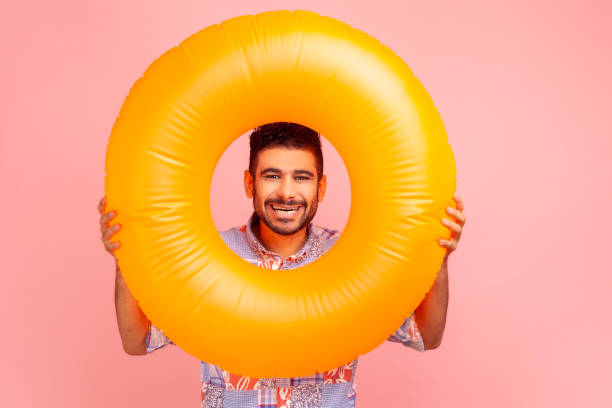 junger erwachsener bärtiger mann mit glücklichem gesichtsausdruck, der durch leuchtend orangefarbenen gummiring schaut, in die kamera schaut und seinen urlaub genießt. - swim ring stock-fotos und bilder