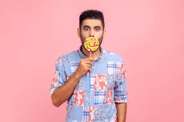 Photo of Young adult bearded man wearing blue casual shirt covering his astonished face with round sweet sugary rainbow candy, looking at camera with big eyes.