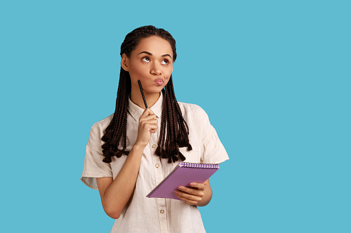 Portrait of pensive woman with black dreadlocks holding paper notebook, having thoughtful facial expression, planning, wearing white shirt. Indoor studio shot isolated on blue background.