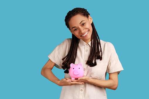 Portrait of happy satisfied woman with black dreadlocks holding piggy bank and looking at camera with toothy smile, money savings, wearing white shirt. Indoor studio shot isolated on blue background.