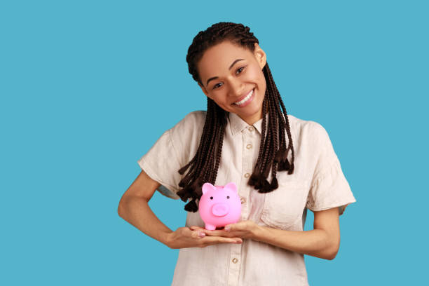 Woman with black dreadlocks holding piggy bank and looking at camera with toothy smile, money saving Portrait of happy satisfied woman with black dreadlocks holding piggy bank and looking at camera with toothy smile, money savings, wearing white shirt. Indoor studio shot isolated on blue background. a penny saved stock pictures, royalty-free photos & images