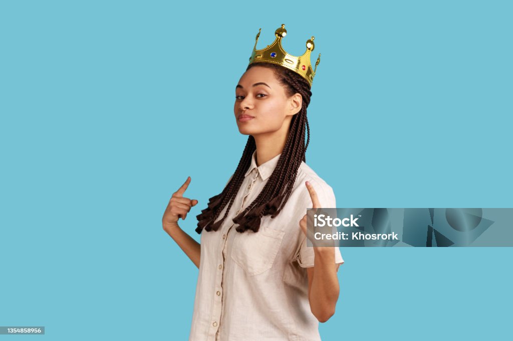 Arrogant woman with dreadlocks, standing in gold crown, pointing at herself with pride. Portrait of proud confident arrogant woman with black dreadlocks, standing in gold crown, pointing at herself with pride, wearing white shirt. Indoor studio shot isolated on blue background. Showing Off Stock Photo