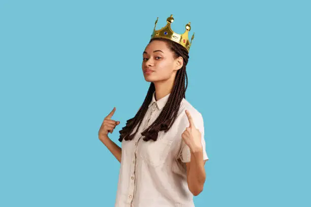 Photo of Arrogant woman with dreadlocks, standing in gold crown, pointing at herself with pride.