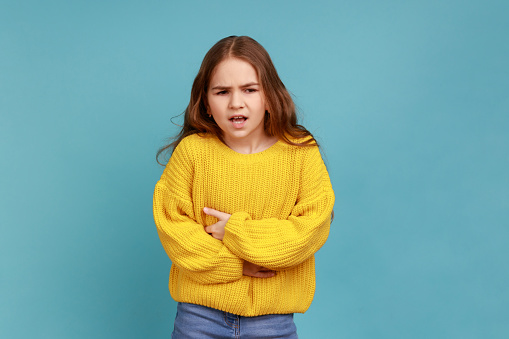 Little girl clutching belly, feels discomfort or pain in stomach, suffering constipation cramps, wearing yellow casual style sweater. Indoor studio shot isolated on blue background.