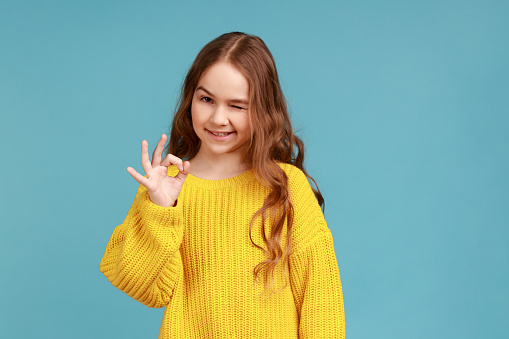Portrait of adorable little girl showing ok gesture to camera and winking, expression optimism, wearing yellow casual style sweater. Indoor studio shot isolated on blue background.