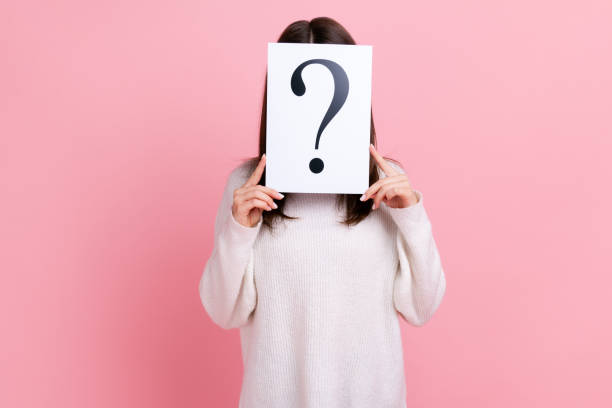 Unknown shy female standing hiding face behind paper with question mark, having problem for solving.