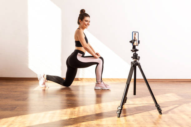 istock Online trainer female stretching leg while broadcasting livestream, using phone camera on tripod. 1354865155