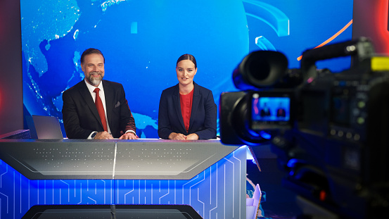 Live News Studio with Beautiful Female and Handsome Man Anchors Start Reporting. TV Broadcasting Channel with Presenters Talking. Television Newsroom Set. Behind the Scene Camera Shooting Shot