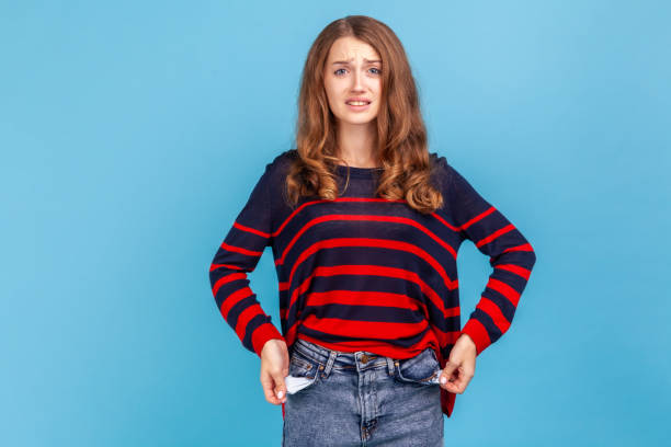 Woman turning empty pockets inside out, looking frustrated by overspend, lack of money. I'm bankrupt! Unhappy poor woman, turning empty pockets inside out and looking frustrated by overspend, lack of money, wearing striped casual sweater. Indoor studio shot isolated on blue background. over spend stock pictures, royalty-free photos & images