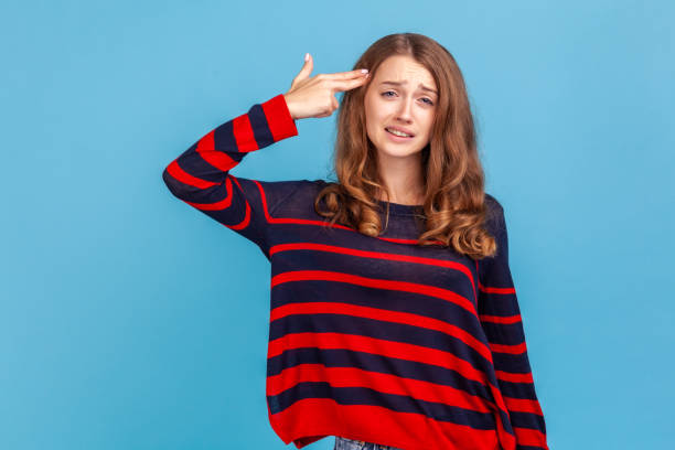 Woman pointing finger gun to head, shooting herself, making suicide gesture to stop depression. Kill me please! Portrait of desperate woman, pointing finger gun to head, shooting herself, making suicide gesture to stop depression, wearing sweater. Indoor studio shot isolated on blue background. head shoot stock pictures, royalty-free photos & images