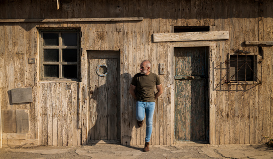 Portrait of adult man in jeans standing against wood wall with doors in summer