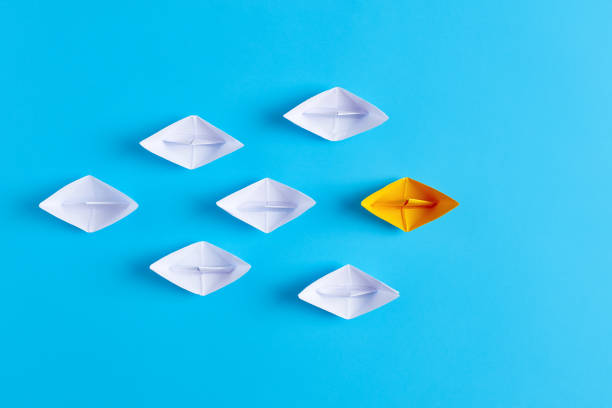 istock Yellow paper boat leads white paper ships. Leadership, collaboration, cooperation, or partnership in business 1354878465