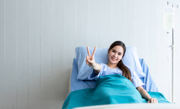 asiatische junge patientin smiley gesicht hebt zwei finger hoch kämpfen mit krankheit auf dem bett im zimmer krankenhaus hintergrund. - patient room stock-fotos und bilder
