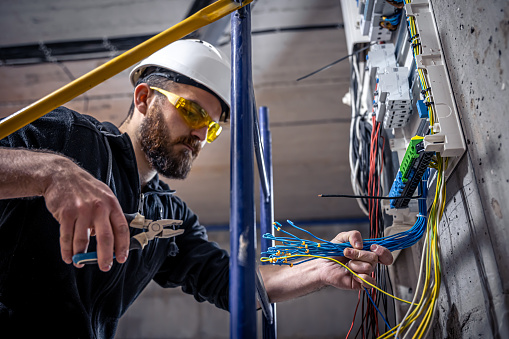 A male electrician works in a switchboard with an electrical connecting cable, connects the equipment with tools.