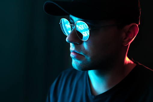 Man with neon light on glasses and face. Blue and red color from city billboard in dark studio portrait. Reflection in sunglasses. Black background in dramatic urban night portrait. Cyber technology.