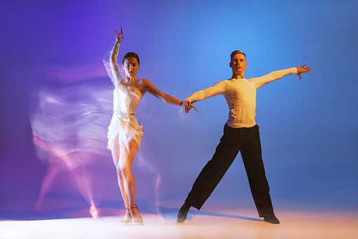 Retrato dinámico de bailarines elegantes, hombres y mujeres flexibles bailando baile de salón aislado sobre fondo púrpura azul degradado en luz mixta de neón photo