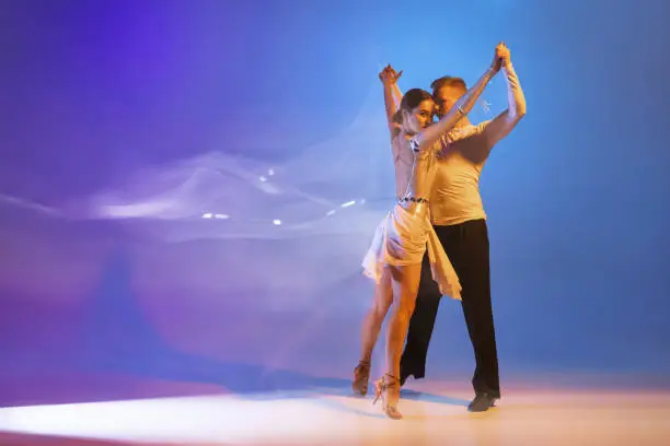 Photo of Two young graceful dancers, flexible man and woman dancing ballroom dance isolated on gradient blue purple background in neon mixed light