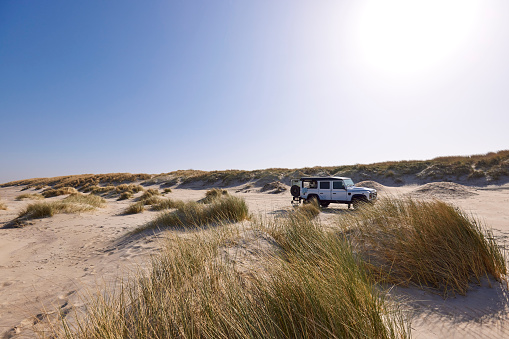 A white Landrover Defender 110 4X4 car on the beach in Zeeland during springtime. The sky is blue and the sun is shining brightly. The grass is standing tall on the sand dunes.