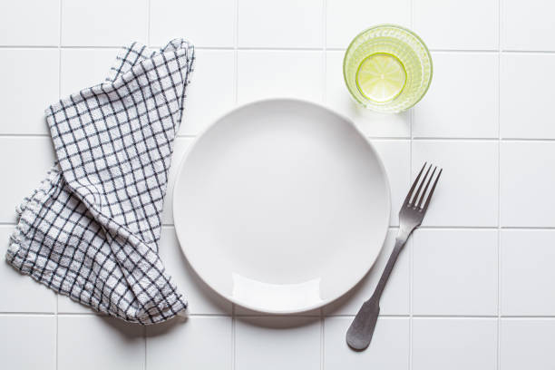 Empty white plate with fork, napkin and glass of water on white tile background, copy space, top view. Intermittent fasting, healthy lifestyle, diet concept.