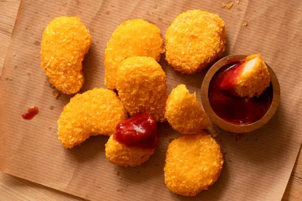 Fried gluten free cornflake crumb chicken nuggets next to a wood bowl of ketchup on brown baking paper. Dipped nugget. Top view.