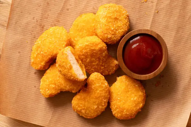 Fried gluten free cornflake crumb chicken nuggets next to a wood bowl of ketchup on brown baking paper. Top view.