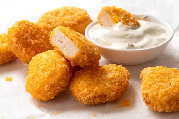 Fried gluten free cornflake crumb chicken nuggets next to a bowl of white sauce on white baking paper. Dipped nugget.