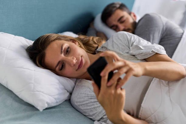 istock Young woman using mobile phone and checking messages in bed while her husband is asleep 1354907880