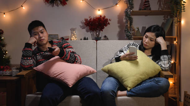 bored japanese man propping face and surfing channels with a tv control while his wife is using phone, ignoring each other on the sofa on christmas night in the living room at home - gamer watching tv adult couple imagens e fotografias de stock