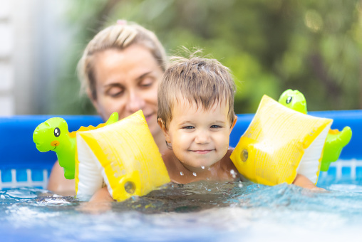 Happy mom playing with little laughing naked baby with colorful red inflatable armbands, in pool with clear clean cool water against backdrop of bright golden summer sunny sunset