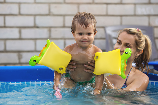 Happy mom playing with little laughing naked baby with colorful red inflatable armbands, in pool with clear clean cool water against backdrop of bright golden summer sunny sunset