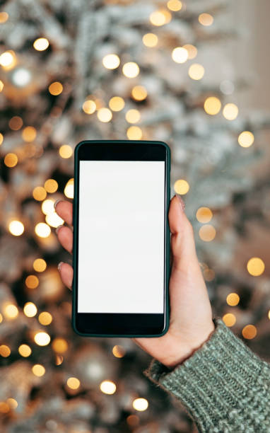 istock Close-up smartphone with empty screen mockup on holiday background with Christmas tree, customer holding phone in hand 1354916789