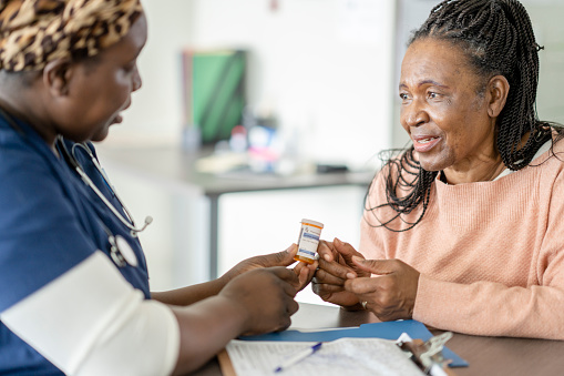 A senior woman sits across the desk from a nurse practitioner as they discuss her medications.  She is dressed casually and looking at the nurse as she listens attentively.  The nurse is dressed in blue scrubs and has a headscarf on as she holds out the medication and informs the woman about how to take the new medication.