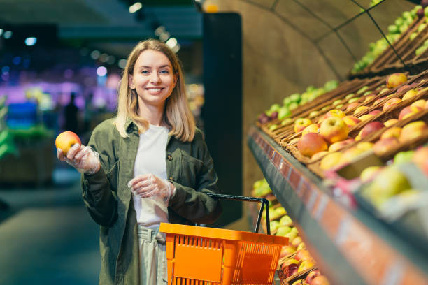 若いブロンドの女性は、スーパーマーケットのカウンターで果物の野菜を選びます。野菜デパートの近くに立ち、バスケットを手にして市場で買い物をする女性主婦。りんごを調べる - vegies vegetable basket residential structure ストックフォトと画像
