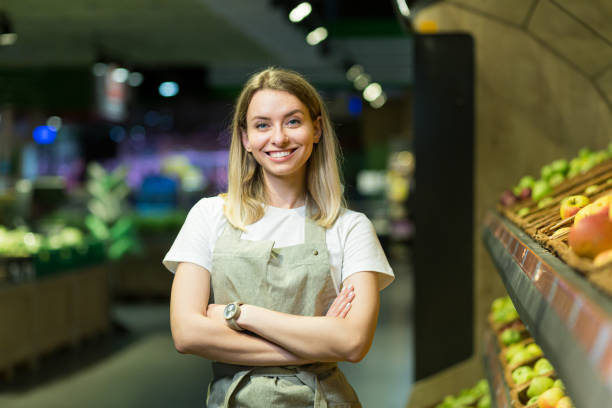 Portrait young woman worker seller in a Vegetable section supermarket standing in arms crossed. greengrocer female looking at camera in fruit shop market Employee in a work apron Portrait young woman worker seller in a Vegetable section supermarket standing in arms crossed. greengrocer female looking at camera in fruit shop market Employee in a work apron market vendor stock pictures, royalty-free photos & images