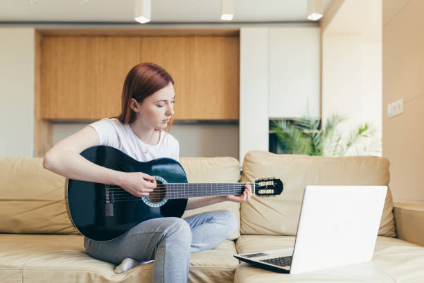 istock young red haired girl learns to play the guitar with the help of video tutorial lessons. female woman home at leisure sitting on sofa with laptop studying online musical instrument. Hobbies distance 1354939672