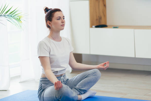 young attractive woman is practicing yoga online using a mobile phone or smartphone app. Female yogi morning in casual clothes at home in living room, workout lotus position meditates on a mat una mujer joven y atractiva está practicando yoga en línea usando un teléfono móvil o una aplicación para teléfonos inteligentes. mujer yogui mañana con ropa casual en casa en la sala de estar, entrenamiento posición de loto medita en una colchonet