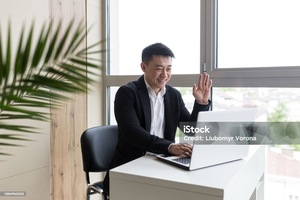 Successful Asian businessman, in a black business suit, works on a laptop, in a stylish office explains a new business strategy via video link Asia Stock Photo
